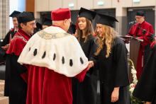 A person in a black academic gown and mortarboard shakes hands with a person in ceremonial red-and-white academic attire during the diploma presentation ceremony.