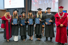 Seven people pose on stage during the ceremony. Five graduates in black academic gowns and mortarboards hold navy-blue folders, while two people in red academic robes stand on either side.
