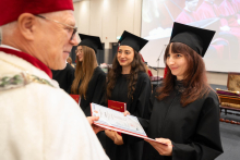 A person in a black academic gown and mortarboard receives a diploma from a person in ceremonial red-and-white academic attire during the ceremony.
