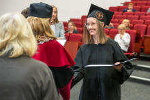 A female graduate in a black academic gown and mortarboard shakes hands with a person in red academic attire during the diploma presentation. Other graduates are waiting in the background.