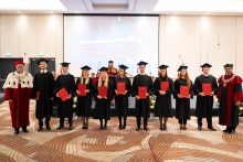 A group of people in black academic gowns and mortarboards pose with red diploma folders between two people wearing ceremonial academic robes.