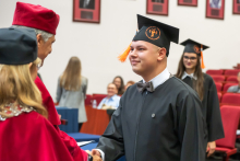 A male graduate in a black academic gown and mortarboard shakes hands with a person in red academic attire during the diploma presentation. Other graduates are waiting in the background.
