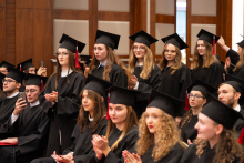 A group of participants in black academic gowns and mortarboards sit and stand in rows during an academic ceremony; some are applauding.