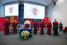 University authorities in academic dress stand on stage during the ceremony. Screens displaying the faculty crest and university banners are visible in the background.