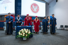 Six people in academic gowns stand in an auditorium during the matriculation ceremony for students of the WUM Faculty of Health Sciences. In front of them is a table with academic insignia and a bouquet of flowers, while the Medical University of Warsaw emblem is displayed on a screen in the background.