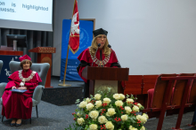 A person wearing a red academic gown, a black mortarboard and a ceremonial chain speaks at a lectern with two microphones. Next to them sits a person wearing a red gown, a red mortarboard and a ceremonial chain. A white floral arrangement is in the foreground.