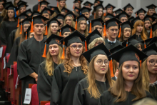 A group of graduates in black academic gowns and mortarboards with orange tassels stand in rows in the auditorium during the ceremony.