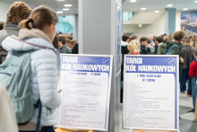 Participants at the Student Research Groups Fair stand by boards listing the participating groups. In the background, a crowd is gathered in the Teaching Centre.