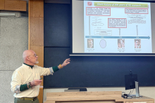 A lecturer is giving a talk in an academic lecture hall and pointing to a slide displayed on the screen. The presentation covers the history of haemoglobin research and includes a timeline and archival photographs of scientists.