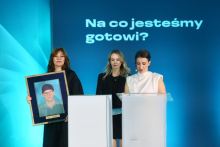 Trzy osoby stoją na scenie podczas wydarzenia. Jedna trzyma oprawiony portret osoby w stroju medycznym, a inna stoi przy mównicy. W tle napis „Na co jesteśmy gotowi?”.