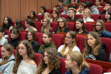 Grupa słuchaczy, głównie młodych kobiet, siedzących w rzędach czerwonych foteli w auli akademickiej i uważnie słuchających.