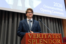 A man speaks at the podium during the opening of Orientation Week 2026. A screen displaying the name of the event is visible in the background.