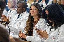 Students wearing white coats are seated in the auditorium during the ceremony. One female student is looking directly at the camera.