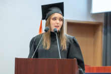 A female graduate in a black academic gown and mortarboard speaks at the lectern.