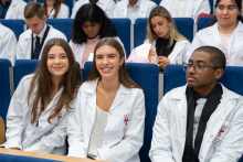 Students wearing white coats are seated in the auditorium during the ceremony. Three people can be seen in the front row. Two of them are smiling for the photo.