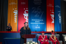 A female representative of the academic community speaks at the lectern during the inauguration of the academic year; members of the university authorities are seated beside her.