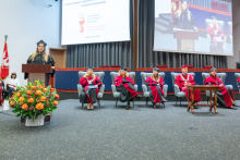 A female student in a black academic gown and mortarboard speaks at the lectern. Six people in academic attire are seated on the stage, and a live feed of the speech is displayed on the screen.