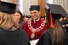 A person in red academic robes with a ceremonial chain stands opposite people in black academic gowns and mortarboards during an academic ceremony.
