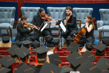 Four people perform a piece on string instruments in front of an audience. Three are playing violins or viola, while one person is playing the cello.