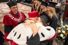 A person in a black academic gown and mortarboard embraces a person in ceremonial red-and-white academic attire during the diploma presentation ceremony.