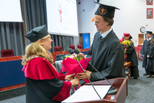 A male graduate presents a flower to a woman wearing an academic gown. Other people can be seen in the background.