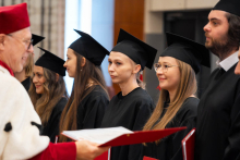 A group of people in black academic gowns and mortarboards stand in line during the diploma presentation ceremony; a diploma is being presented in the foreground.