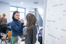 Stylistka marki Mary Kay wykonująca makijaż próbny na twarzy studentki siedzącej przy stanowisku kosmetycznym w pobliżu ścianki z serduszkami.