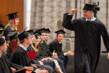 A person in a black academic gown and mortarboard walks past seated participants in an academic ceremony, raising a hand in greeting.
