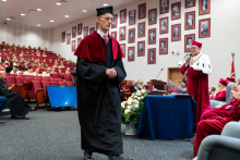 A person wearing an academic gown walks through the auditorium during the ceremony while a speech is being delivered in the background.