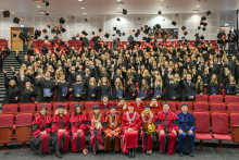 Graduates stand in rows and throw their mortarboards into the air. University representatives are seated in the front rows.