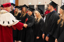 A person in ceremonial red-and-white academic attire shakes hands with one of the people standing in a row in black academic gowns and mortarboards.