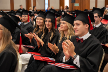 Participants in black academic gowns and mortarboards sit in a row and applaud during an academic ceremony.