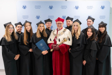 A group of students wearing academic gowns pose for a group photograph with a representative of the university authorities.