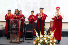 A group of people in red academic robes stand on stage and applaud; in front of them is a lectern bearing the words “Medical University of Warsaw” and a floral arrangement.
