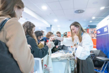 Uśmiechnięta wystawczyni w białej koszuli wręcza materiały promocyjne i torbę z logo studentce stojącej przy stoisku informacyjnym.