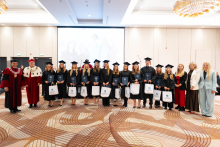 A group of people pose for a group photo during an academic ceremony; people in black academic gowns and mortarboards hold diplomas and gift bags.