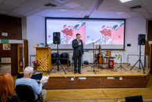 Mężczyzna stoi na scenie i przemawia do uczestników konferencji. Za nim znajdują się ekrany z grafiką wydarzenia oraz instrumenty muzyczne.