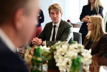 Several people attend a meeting at a conference table. Two people are seated next to each other and taking part in a discussion. Bottles of water and an arrangement of white flowers are on the table.
