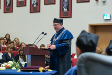 A person wearing a blue academic gown and ceremonial chain speaks at a lectern in an auditorium. Ceremony participants are seated in the background, with framed photographs displayed on the wall.