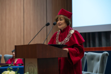 A person wearing a red academic gown, a red mortarboard and a ceremonial chain speaks at a lectern with two microphones, holding a black folder in front of them. Chairs and part of a screen are in the background.