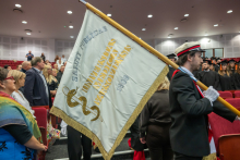 A person in ceremonial attire carries a banner bearing the words “Saluti Publicae” and the name of the Medical University of Warsaw. In the background, ceremony attendees are standing in the auditorium.