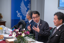 Three people are seated at a conference table during a meeting. One person is speaking and gesturing. In the background, a blue roll-up banner with the WUM logo is visible.