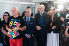 Several people are standing in a clinical room during a media event. In the foreground, one person is holding a WOŚP publication. Other participants are standing nearby, and a television camera can be seen on the right.