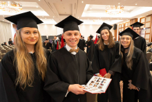 A group of five people in black academic gowns and mortarboards pose in the hall during an academic ceremony; the person in front holds an open photo album.