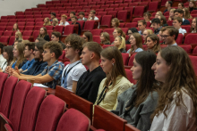 Participants are seated in the auditorium and listen to the presentation. They occupy several rows of red seats in the lecture hall.