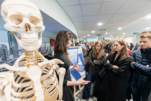 Participants at the Student Research Groups Fair stand by boards listing the participating groups. In the background, a crowd is gathered in the Teaching Centre.