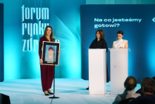 Osoba stojąca przy mikrofonie prezentuje oprawiony portret osoby w stroju medycznym. Obok dwie osoby stoją przy mównicach, a w tle widoczny jest napis „Na co jesteśmy gotowi?”.