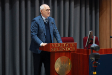 Mężczyzna w okularach i marynarce w kratę stoi za drewnianą mównicą z napisem „VERITATIS SPLENDOR” i przemawia do zgromadzonych