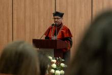 Osoba w czerwono-czarnej todze akademickiej i birecie przemawia przy mównicy wyposażonej w dwa mikrofony.