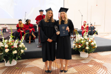Two people in black academic gowns and mortarboards pose during an academic ceremony; one holds a statuette and the other a diploma in a navy-blue cover.
